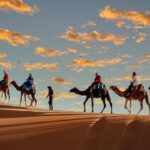 Silhouettes of people riding camels across a sand dune in the Moroccan desert at sunset, with a colorful sky and scattered clouds in the background.
