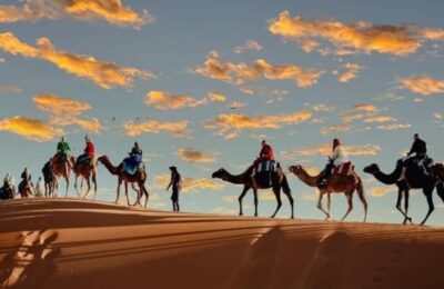 Silhouettes of people riding camels across a sand dune in the Moroccan desert at sunset, with a colorful sky and scattered clouds in the background.