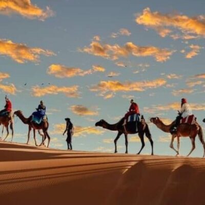 Silhouettes of people riding camels across a sand dune in the Moroccan desert at sunset, with a colorful sky and scattered clouds in the background.
