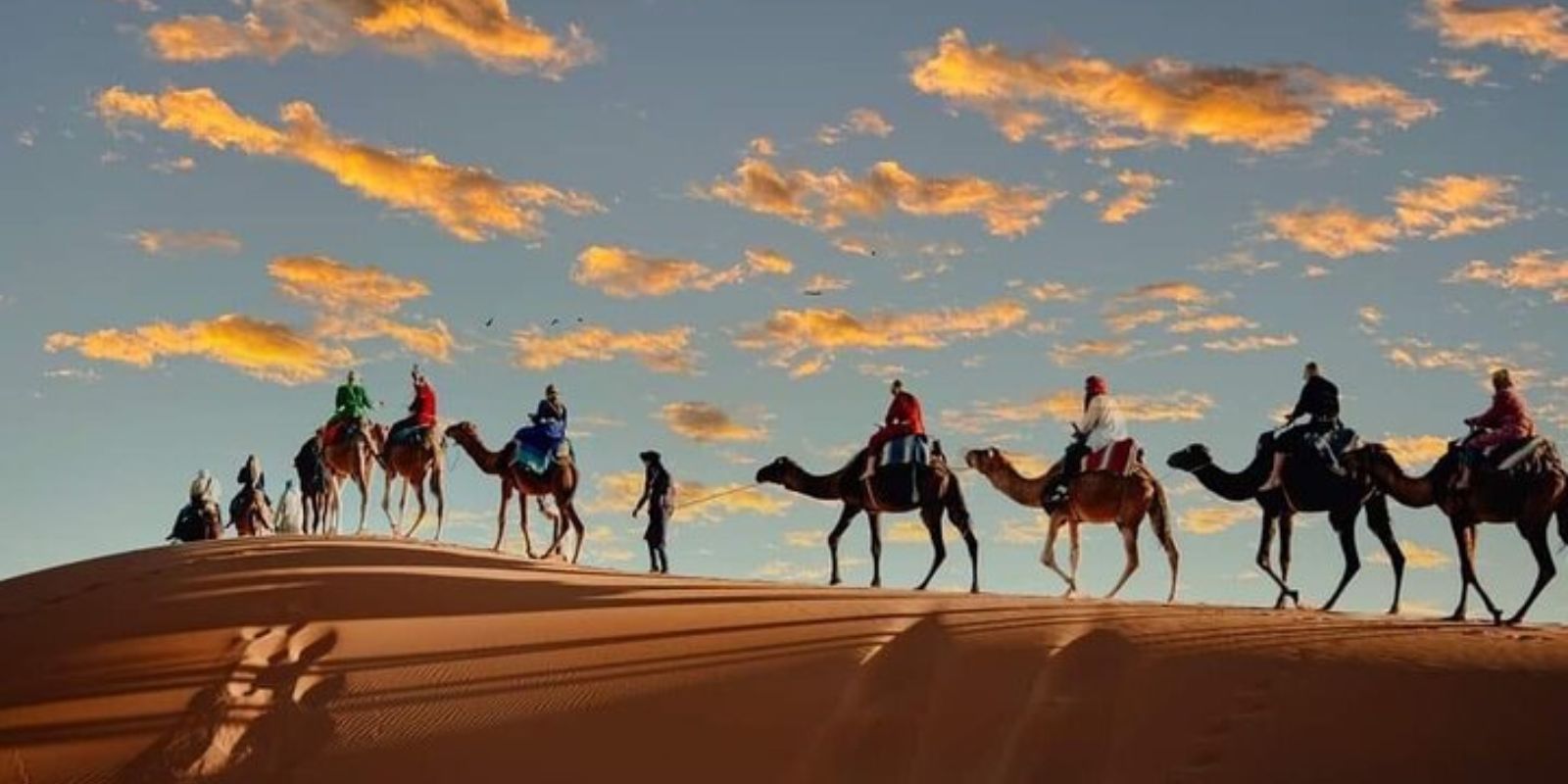 Silhouettes of people riding camels across a sand dune in the Moroccan desert at sunset, with a colorful sky and scattered clouds in the background.