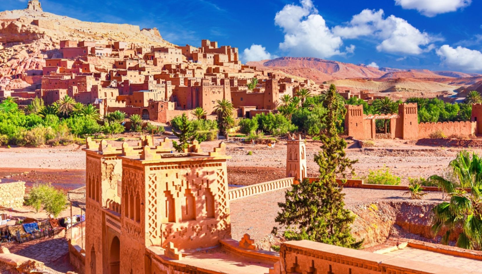 Aït Ben Haddou Kasbah near Ouarzazate, Morocco, with traditional earthen architecture, palm trees, and desert mountains.