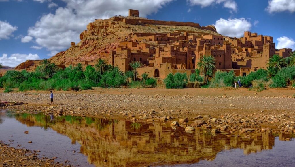 Aït Ben Haddou Ksar near Ouarzazate, Morocco, with its ancient earthen buildings reflected in the water under a bright blue sky.