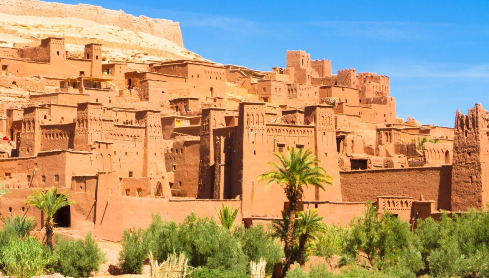 View of Aït Ben Haddou Kasbah near Ouarzazate, Morocco, with traditional clay architecture and palm trees under a bright blue sky.
