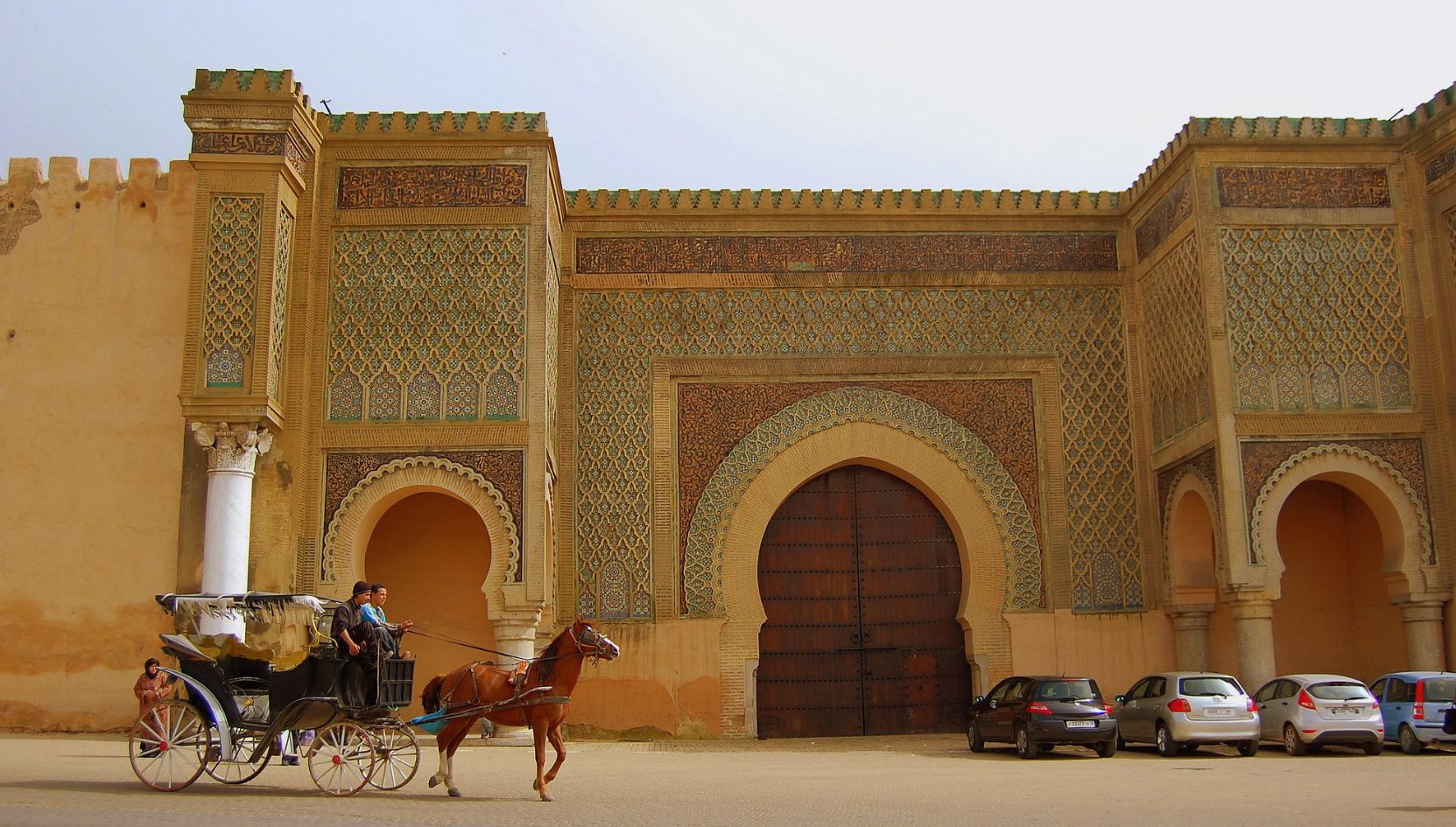Bab Mansour Gate in Meknes, Morocco, with traditional Moroccan mosaic tiles and a horse carriage passing in front.