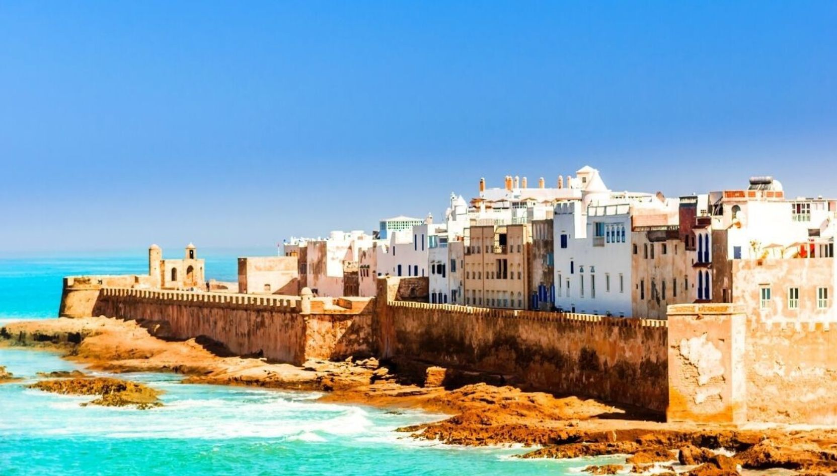 View of Essaouira’s historic ramparts and whitewashed buildings along the Atlantic Ocean under a clear blue sky in Morocco.