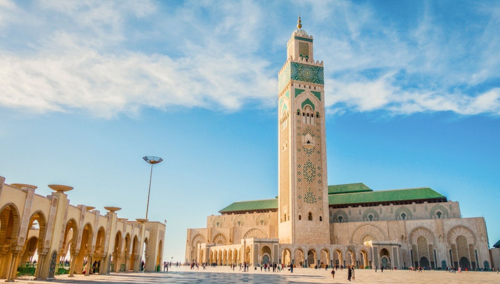 Daytime view of the Hassan II Mosque in Casablanca, Morocco, with clear blue sky and visitors in the courtyard.