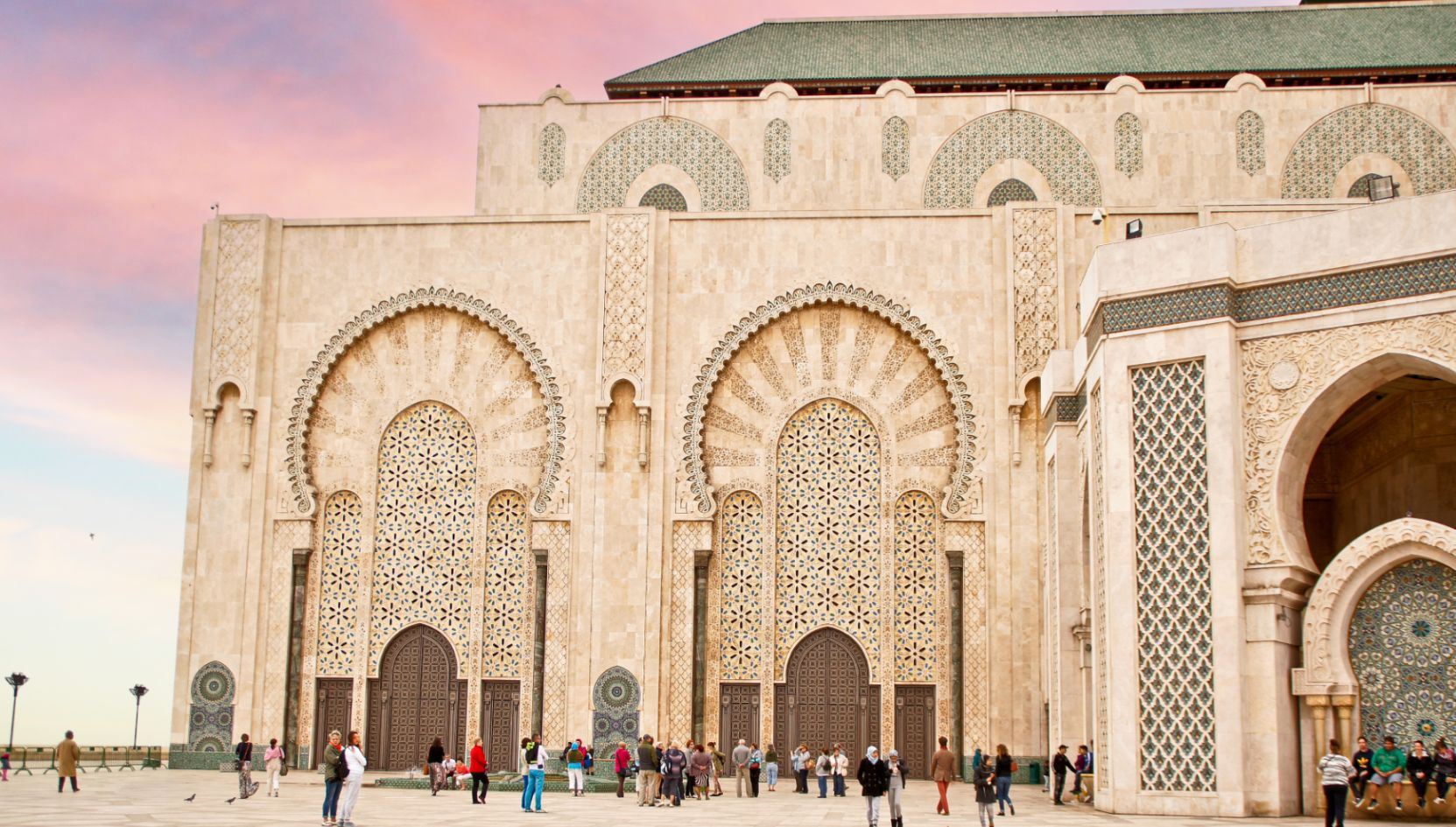 Close-up view of the Hassan II Mosque entrance in Casablanca, Morocco, with intricate Moroccan tilework and people visiting at sunset.