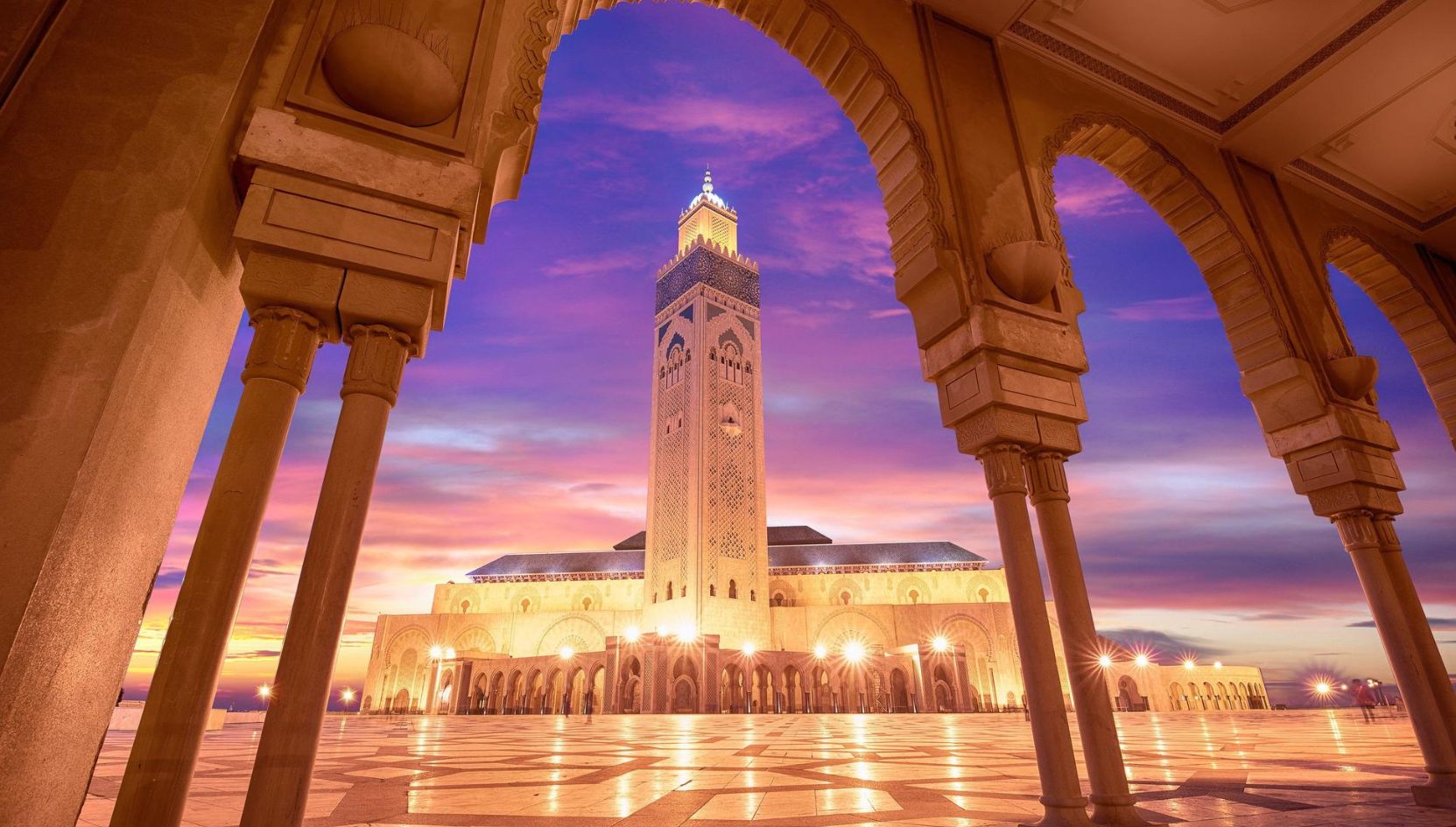 Sunset view of the Hassan II Mosque in Casablanca, Morocco, framed by arches with a colorful sky.