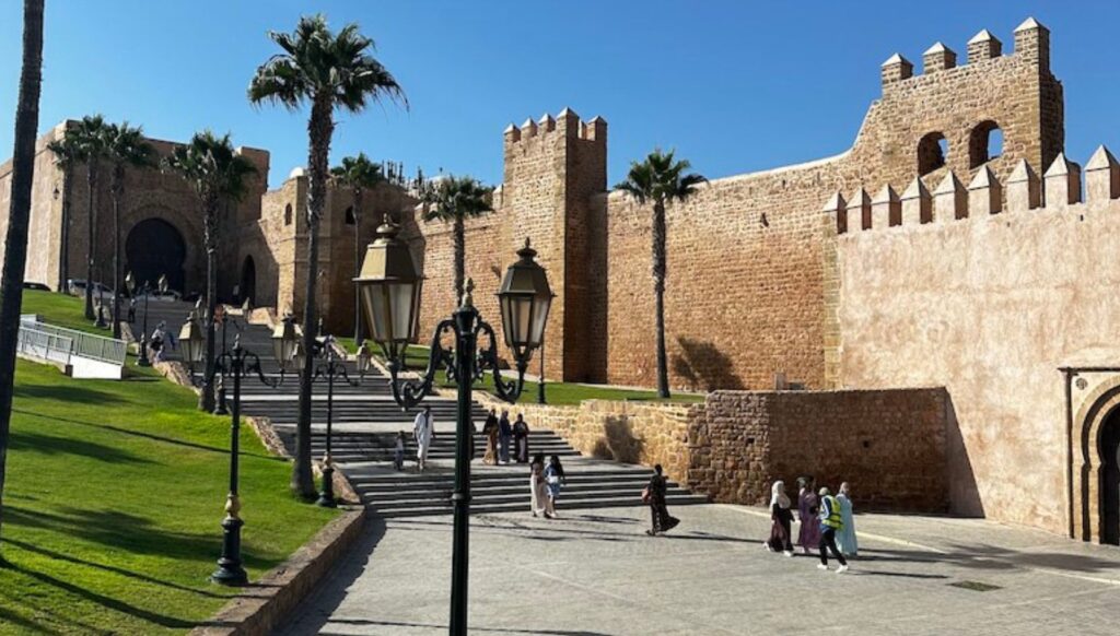 Historic kasbah walls of Marrakech with palm trees and visitors walking near Bab Agnaou gate.