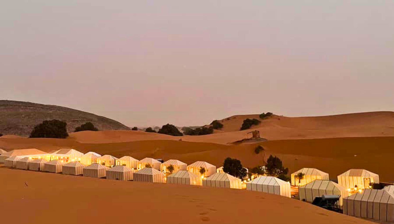 Evening panoramic view of Merzouga Luxury Desert Camp illuminated among the golden dunes of the Sahara Desert.