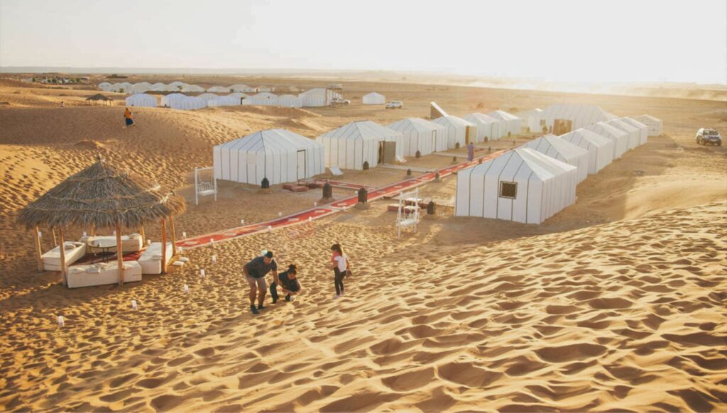Family enjoying the sand dunes near Merzouga Luxury Desert Camp with white tents and a shaded lounge area in the Sahara Desert.
