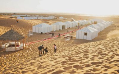 Family enjoying the sand dunes near Merzouga Luxury Desert Camp with white tents and a shaded lounge area in the Sahara Desert.