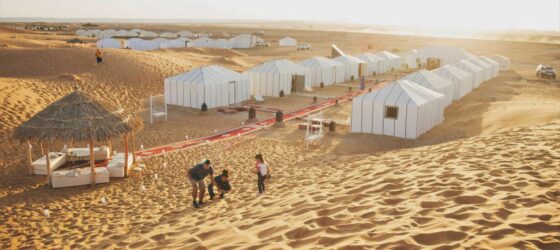 Family enjoying the sand dunes near Merzouga Luxury Desert Camp with white tents and a shaded lounge area in the Sahara Desert.