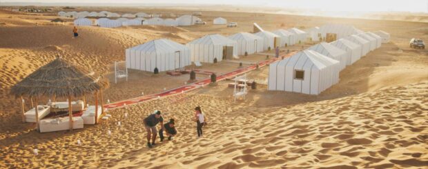 Family enjoying the sand dunes near Merzouga Luxury Desert Camp with white tents and a shaded lounge area in the Sahara Desert.