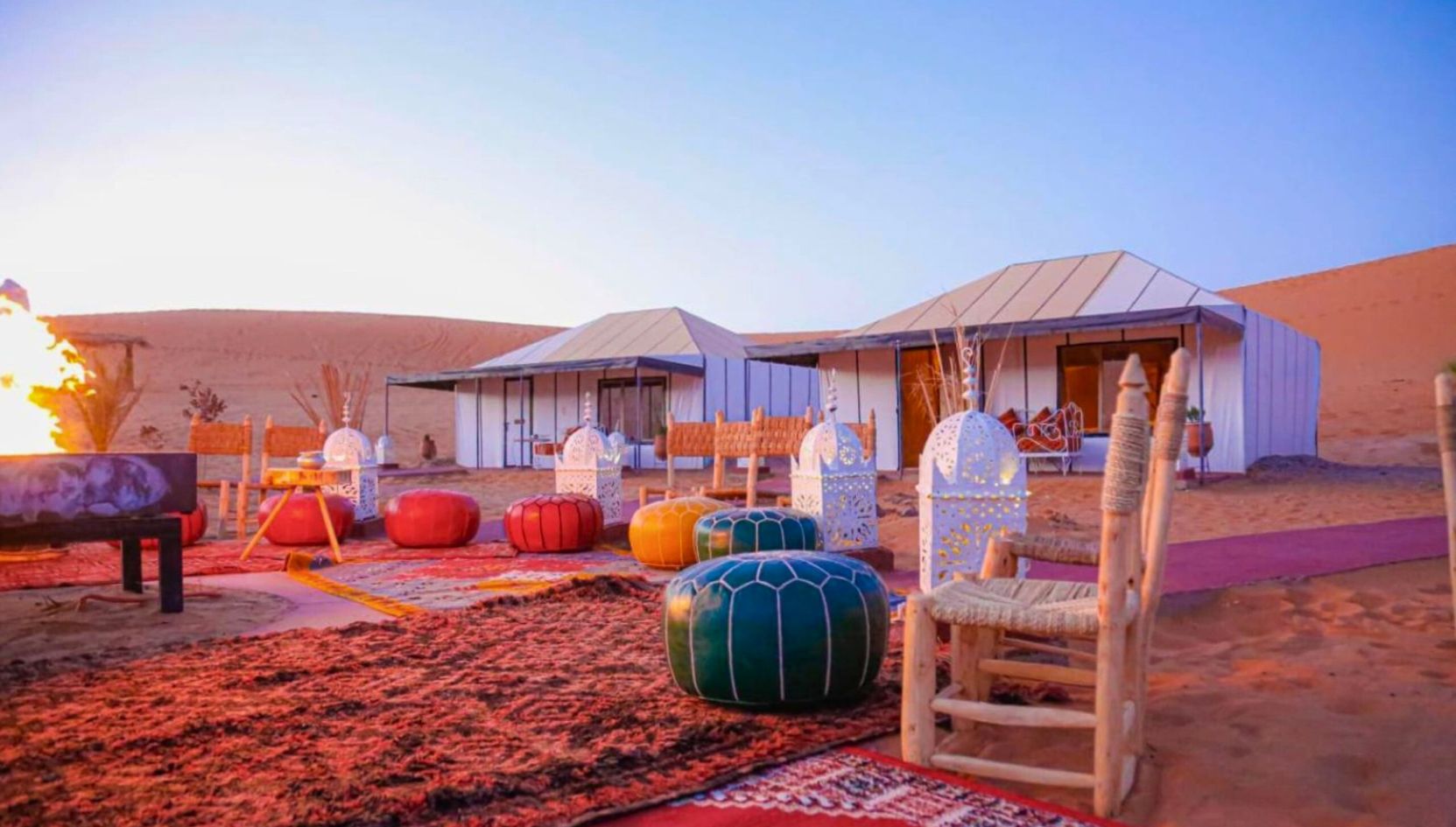 Luxury desert camp in Merzouga with colorful Moroccan poufs, white tents, and evening fire in the Sahara Desert.