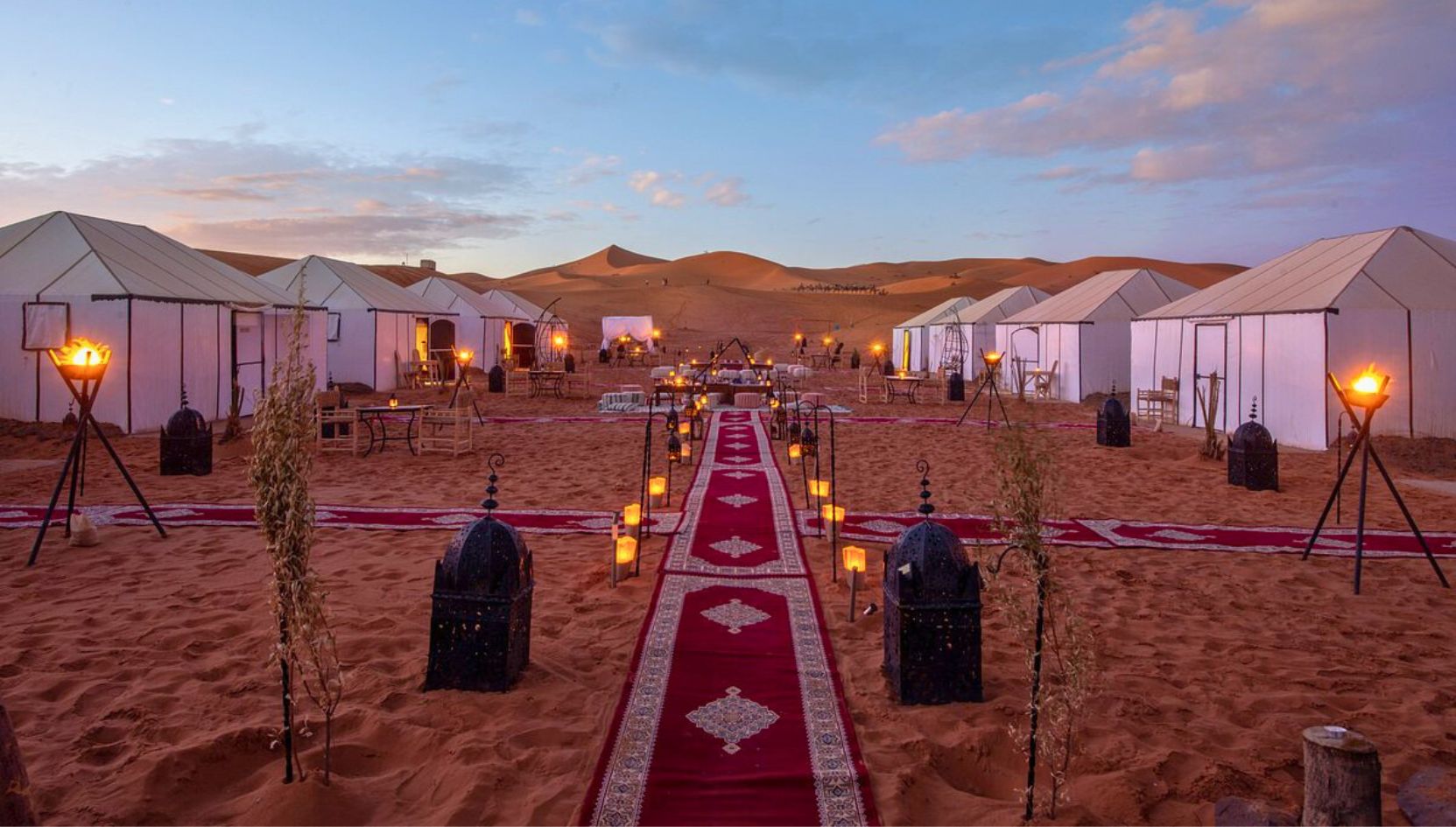 Sunset view of Merzouga Luxury Desert Camp with glowing lanterns, red carpet walkway, and white tents in the Sahara Desert.