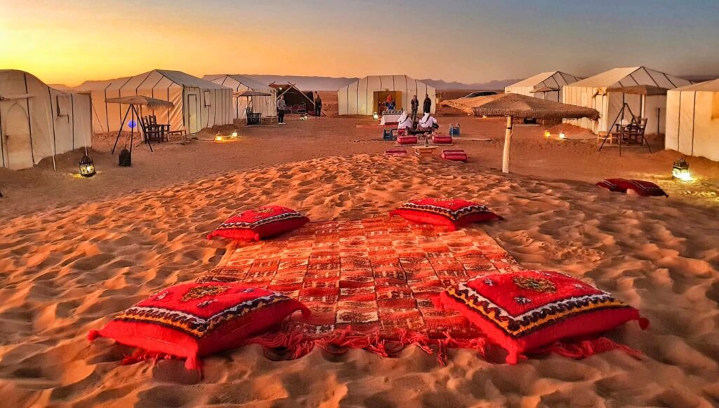 Sunset view of Merzouga Luxury Desert Camp with red Berber cushions and tents illuminated in the golden Sahara dunes.