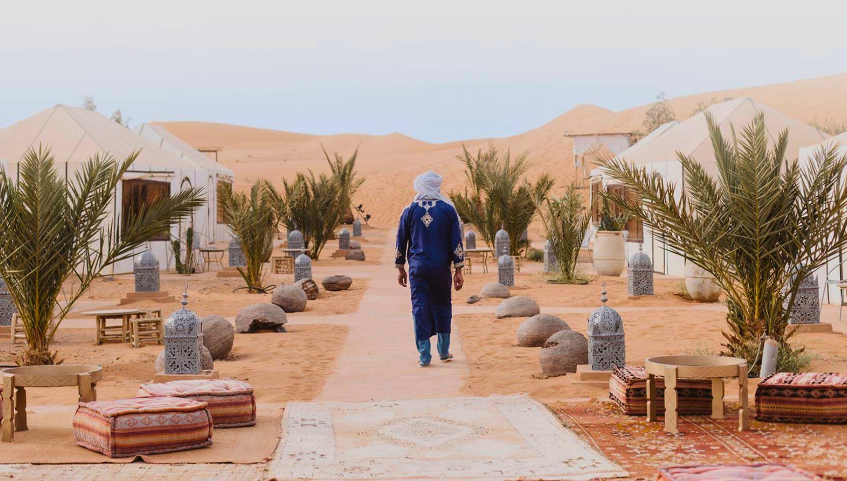 Traditional Moroccan host in blue attire walking through Merzouga Luxury Desert Camp surrounded by palm trees and white tents in the Sahara Desert.