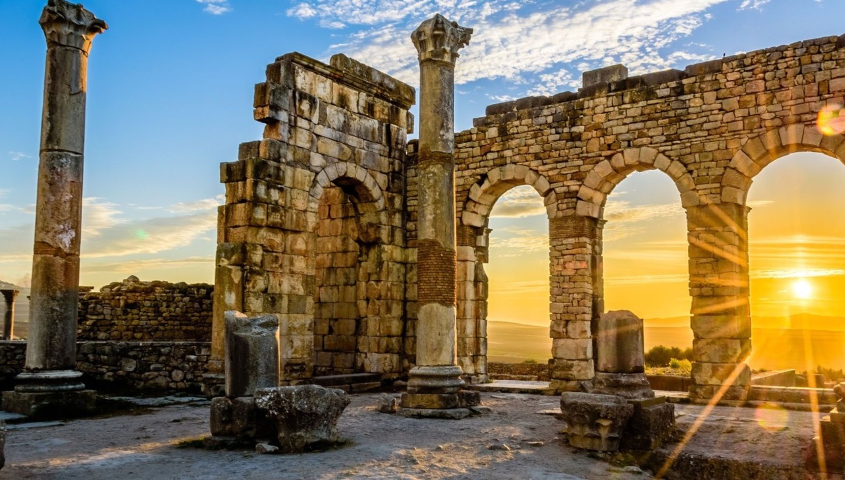 Sunset over the Roman ruins of Volubilis near Meknes, Morocco, with stone arches and ancient columns glowing in golden light.