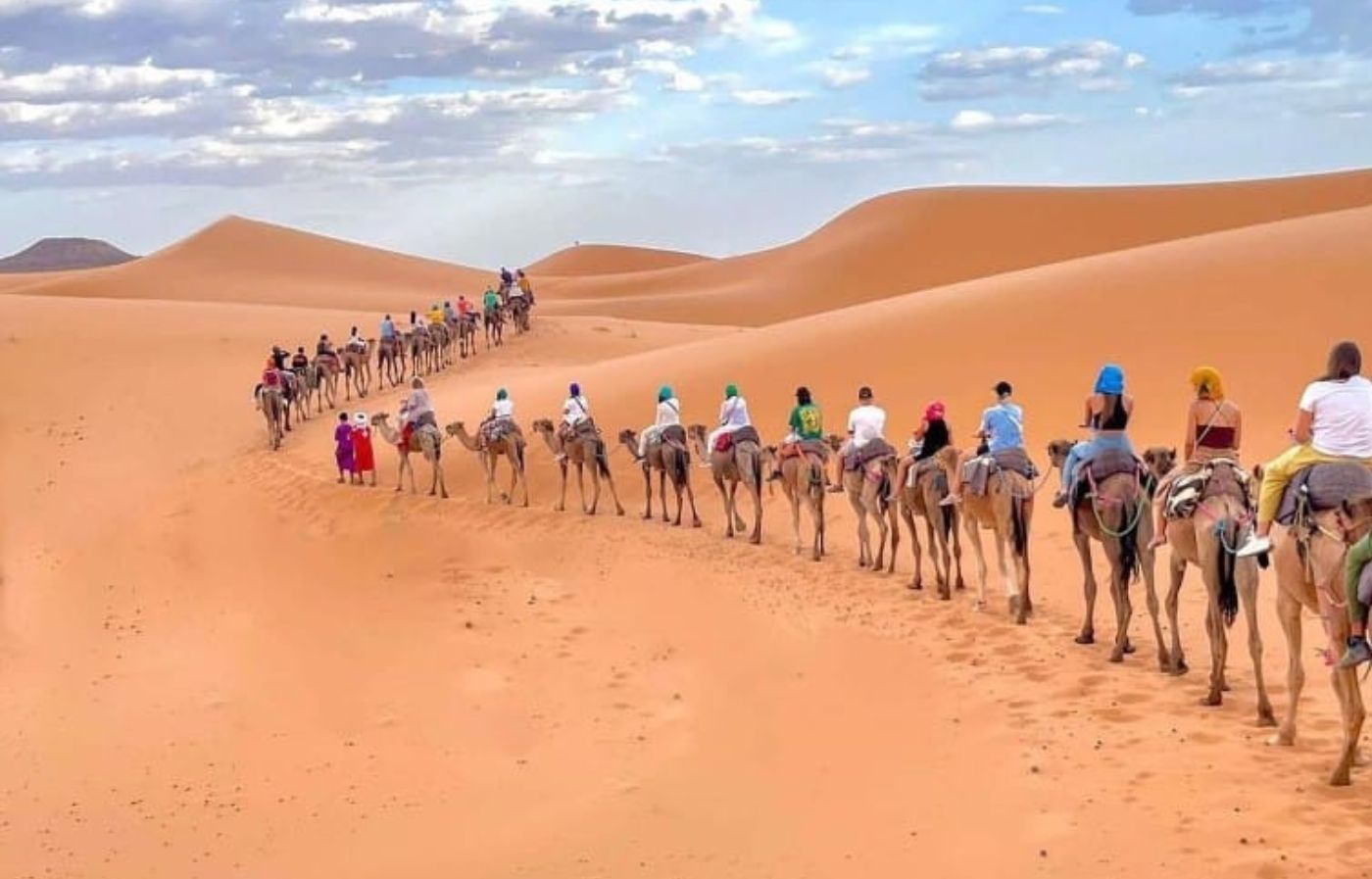 Camel caravan carrying travelers across the golden dunes of the Sahara Desert in Merzouga, Morocco.