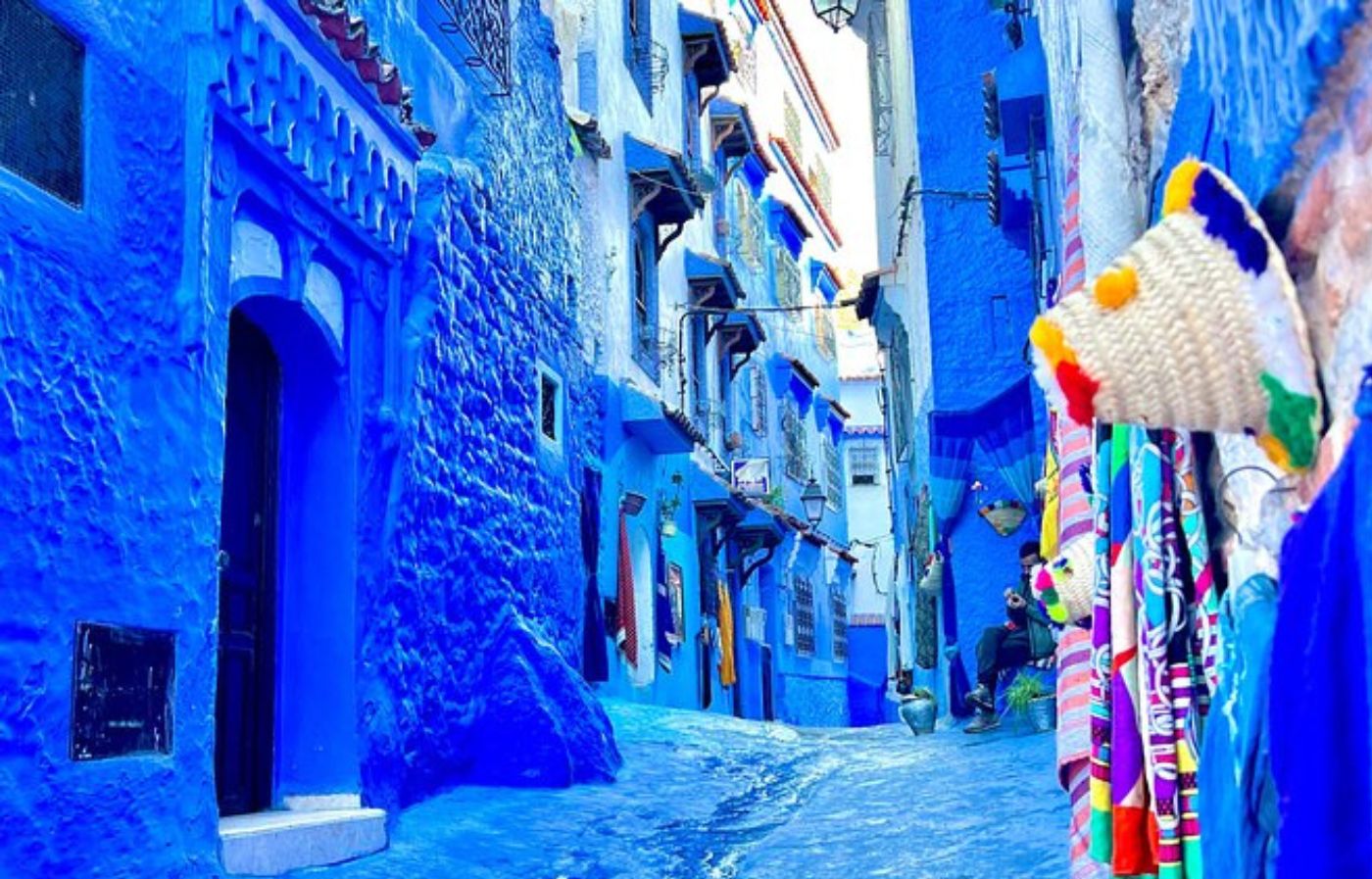 Vibrant blue-painted street in Chefchaouen, Morocco, with traditional houses and colorful local crafts displayed along the walls.