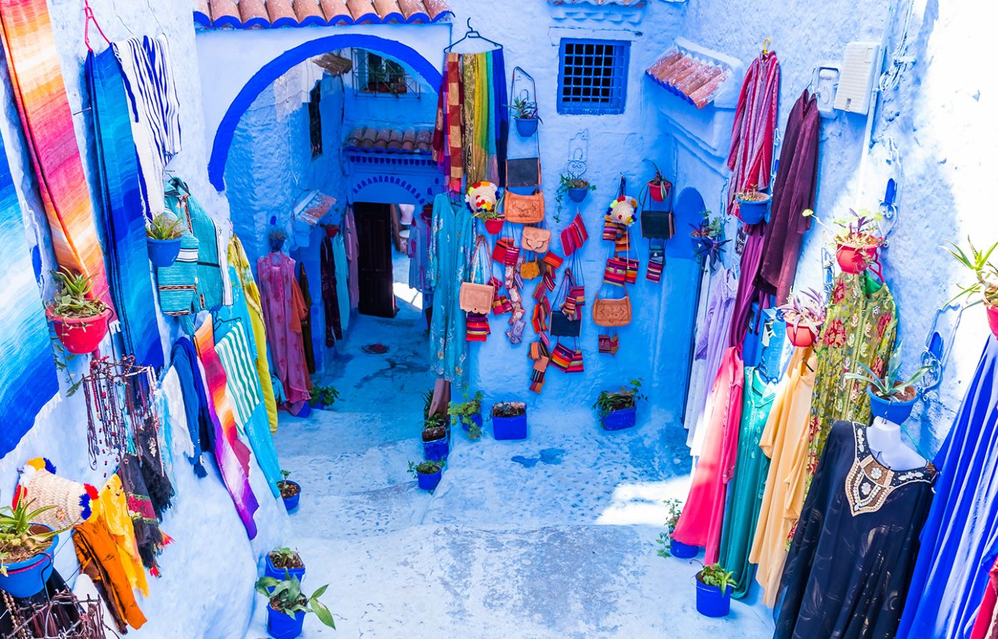 Colorful traditional garments and handicrafts displayed along a blue-painted street in Chefchaouen, Morocco.