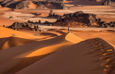 Hikers walking along high Sahara dunes with dramatic rock formations in the Chegaga Desert