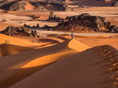 Hikers walking along high Sahara dunes with dramatic rock formations in the Chegaga Desert