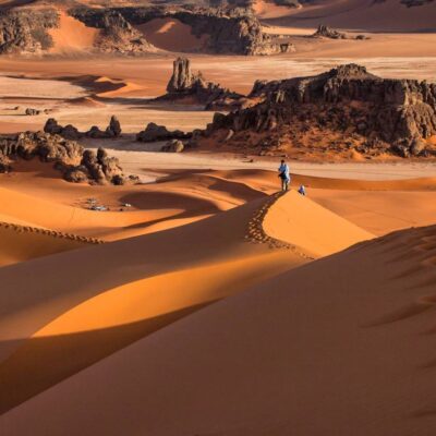 Hikers walking along high Sahara dunes with dramatic rock formations in the Chegaga Desert