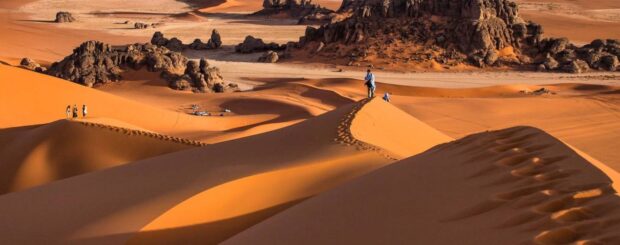 Hikers walking along high Sahara dunes with dramatic rock formations in the Chegaga Desert