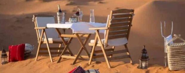 Romantic desert dinner setup with wooden chairs, table, lanterns, and cushions on the sand dunes of Merzouga in Morocco