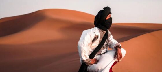 Saharan nomad wearing traditional desert clothing sitting on sand dunes in the Moroccan Sahara.