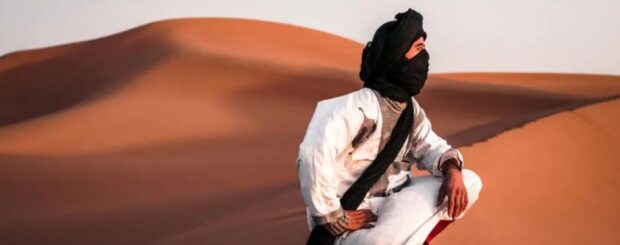 Saharan nomad wearing traditional desert clothing sitting on sand dunes in the Moroccan Sahara.